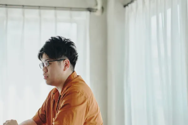 ALTはじまり: 白いカーテンの前で話をしている葉栗さんの横顔。オレンジ色のシャツと黒縁メガネを身につけている。ALTおわり