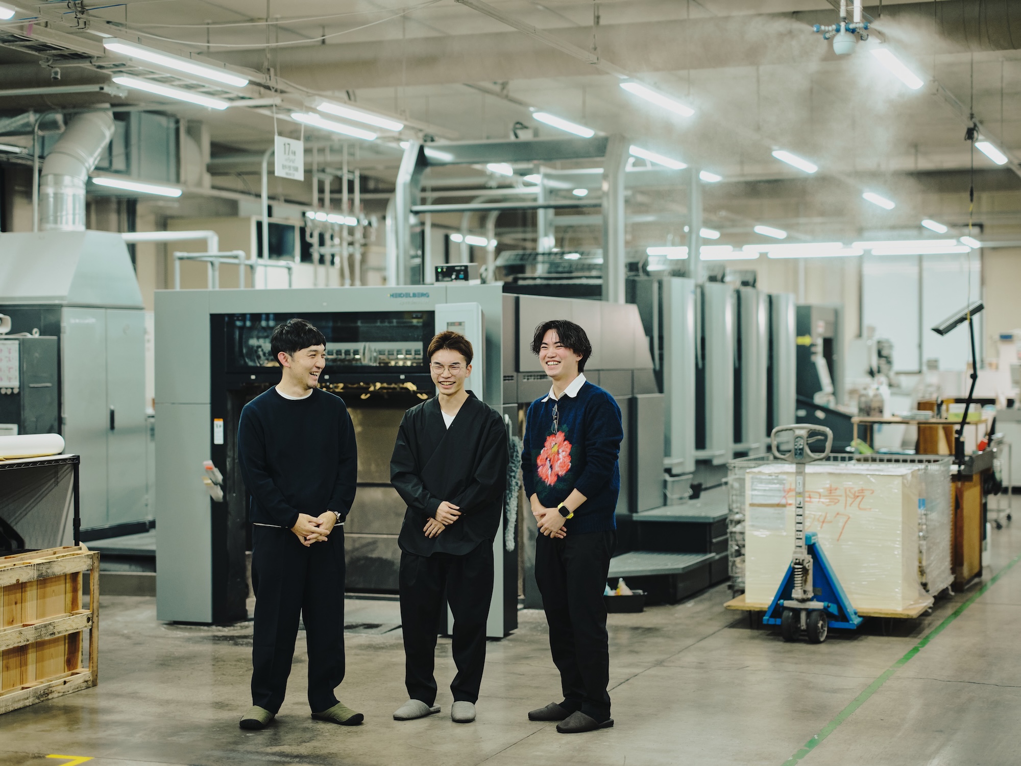 写真:藤原隆充さん、尾木洸太さん、速水駿さんの3名が印刷工場内で並んで立つ。大型印刷機を背景に談笑している。