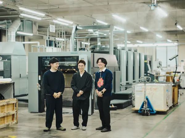 写真:藤原隆充さん、尾木洸太さん、速水駿さんの3名が印刷工場内で並んで立つ。大型印刷機を背景に談笑している。