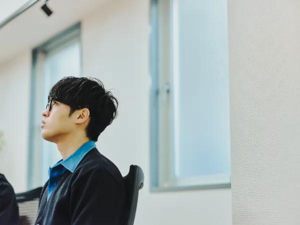  写真：椅子に座り、横を向いて話を聞く松野さん。室内の窓から柔らかな光が入っている。