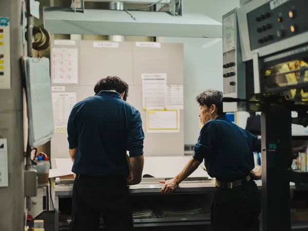写真:藤原印刷の工場で、2人の作業者が刷り上がりの紙を確認している様子。背を向けた若手と横向きのベテランが並び、真剣に仕上がりをチェックしている。