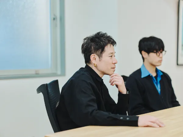 写真：会議室のテーブルに座り、黒いシャツを着た井上さんが顎に手を当てて考え込む様子。隣にブルーのシャツを着た松野さんが座っている。