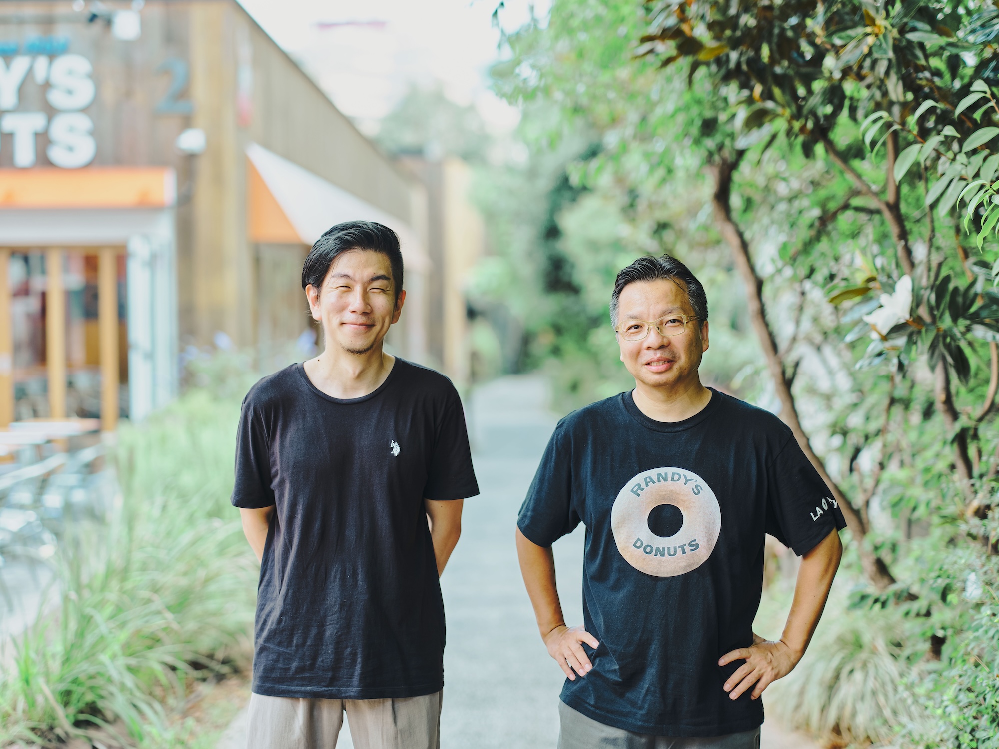 ALTはじまり:東京・代官山にある、「RANDY'S DONUTS」の店先です。木々の緑が映える空間で、芳賀さんと溝口さんが並んでカメラに向かって微笑んでいます。ALTおわり