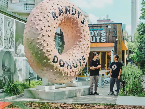 ALTはじまり:ALTテキスト: 東京・代官山の「RANDY'S DONUTS」の前に置かれた、本物そっくりな巨大なドーナツ。芳賀さんと溝口さんが、その遊び心あるオブジェの横で笑顔を見せている。ALTおわり