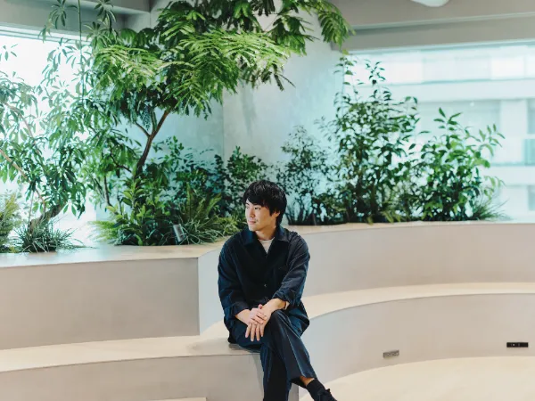 ALTはじまり：植栽に囲まれたスペースで、リラックスした様子で座り、遠くを見つめる兼原さん。場所は、25年9月にオープンしたばかりの丸井グループ本社内にある「共創LOUNGE」。世界的なデザインオフィスnendoが監修した。ALTおわり