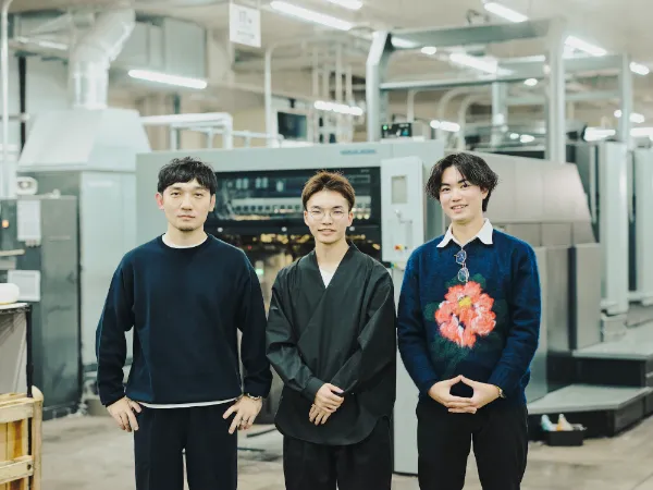 写真:工場の印刷機前で並ぶ藤原隆充さん、尾木洸太さん、速水駿さん。新サイト制作をともに進めた3名のおだやかな表情。