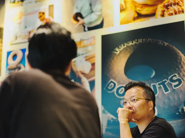 ALTはじまり:隣にいる溝口さんの方向を見ながら、顎に手を当てて話す様子の芳賀さん。背景の大きなポスターには、店頭にある巨大なドーナツのオブジェの写真が飾られています。 ALTおわり