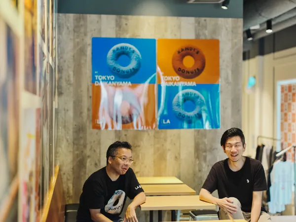 ALTはじまり:店内の大きな窓から光が差し込む、明るい空間です。芳賀さんと溝口さんがテーブルを挟み、談笑しています。壁に飾られたポスターが、和やかな雰囲気を引き立てています。ALTおわり