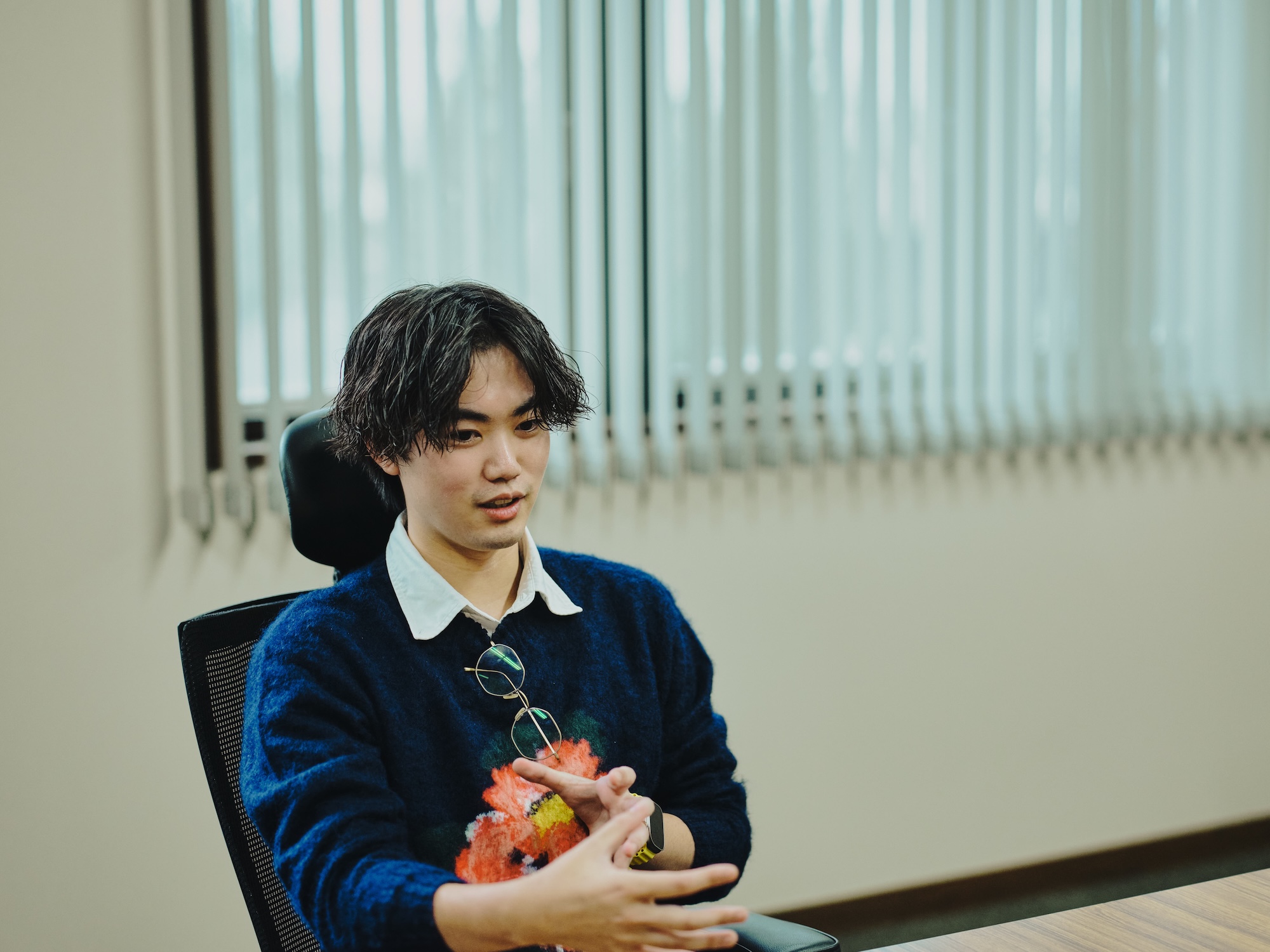 写真:速水さんが手振りを交えて話している様子。紺のセーターを着て、眼鏡を胸元に掛けている。