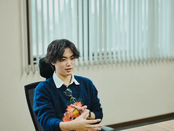 写真:速水さんが手振りを交えて話している様子。紺のセーターを着て、眼鏡を胸元に掛けている。