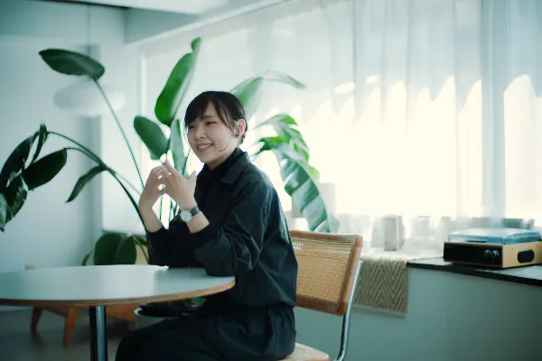  ALTはじまり：テーブルと椅子のある部屋で、笑顔の大橋さんが座っている。背景には大きな観葉植物と窓がある。 ALTおわり