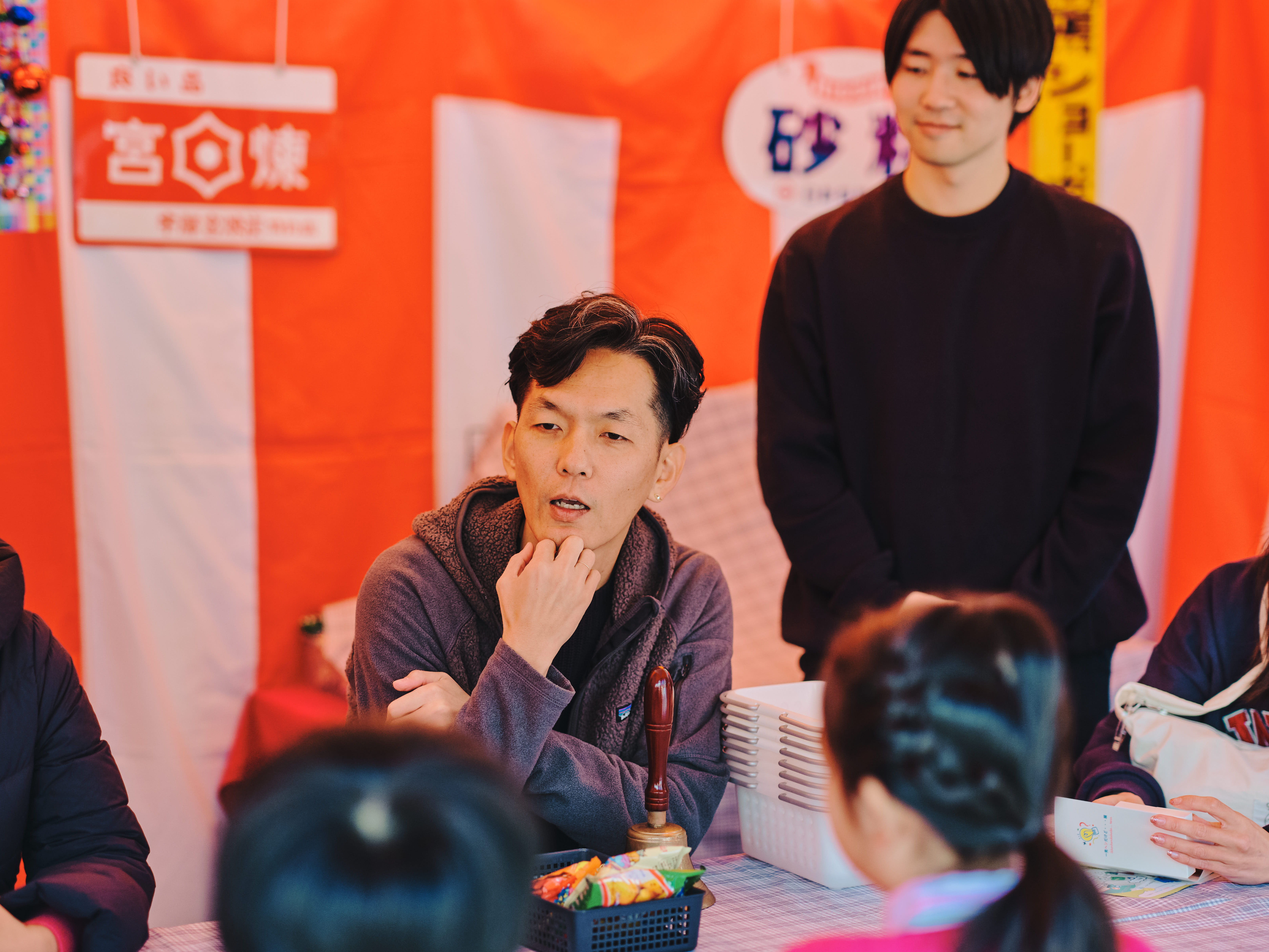 ALTはじまり:赤と白の幕の前に設置された駄菓子屋ブースで、石井さんが子どもたちと向き合って話している。高橋さんは後ろで様子を見守っている。ALTおわり