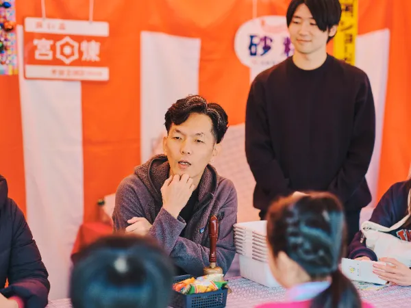 ALTはじまり:赤と白の幕の前に設置された駄菓子屋ブースで、石井さんが子どもたちと向き合って話している。高橋さんは後ろで様子を見守っている。ALTおわり