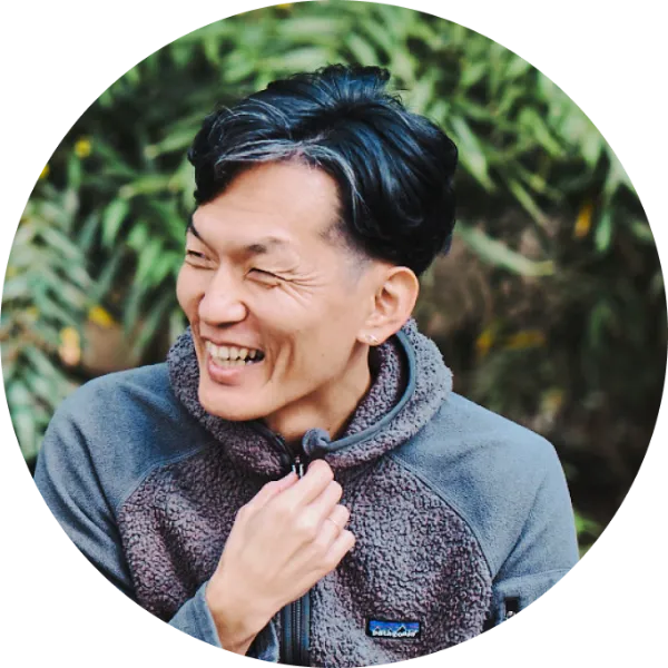 ALTはじまり:屋外で撮影した、石井健介さんのプロフィール写真。笑顔でフリースジャケットの襟元を軽くつまみ、肩の力が抜けた自然な表情をしている。背景には緑が広がっている。ALTおわり