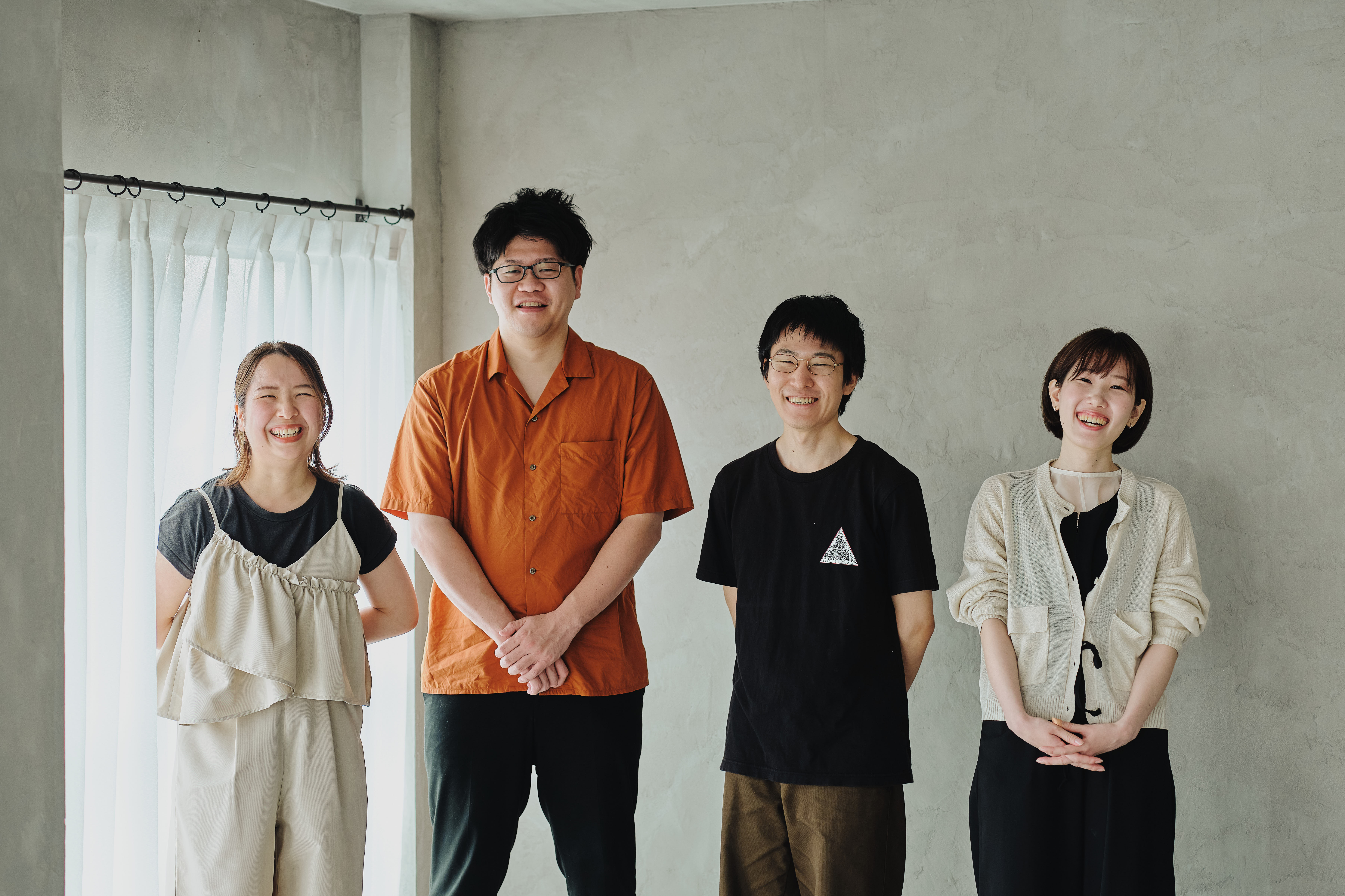 ALTはじまり:白いカーテンとグレーの壁の前で並んで笑顔を見せるcaroaのメンバー4人。左から2番目、オレンジ色のシャツを着た人物が葉栗さん。 ALTおわり