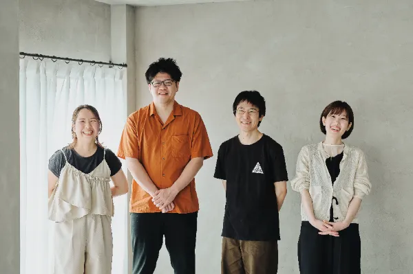 ALTはじまり:白いカーテンとグレーの壁の前で並んで笑顔を見せるcaroaのメンバー4人。左から2番目、オレンジ色のシャツを着た人物が葉栗さん。 ALTおわり