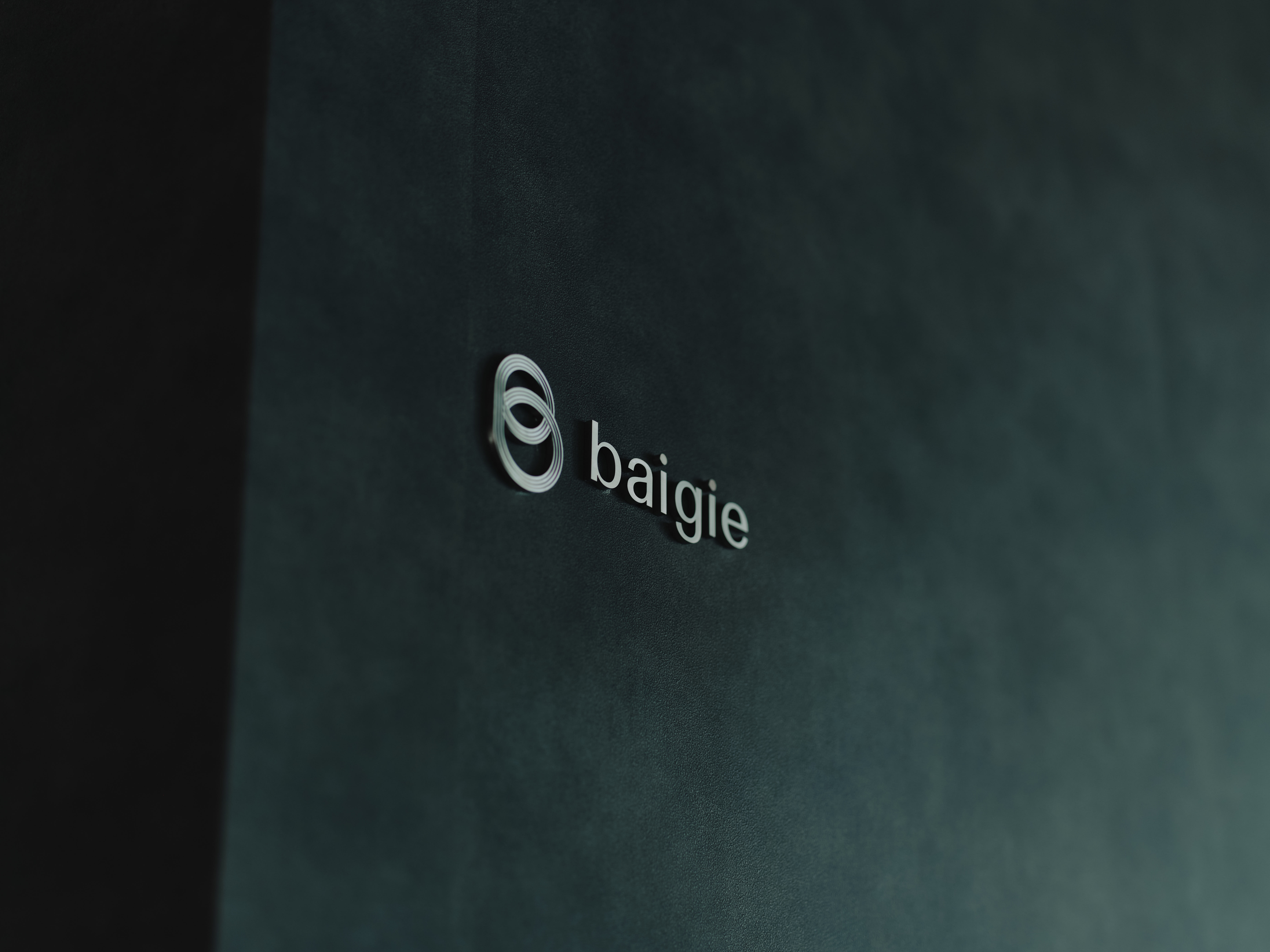 ALTはじまり: ベイジのオフィスにある、黒い背景の壁に会社のロゴ「baigie」が銀色の立体文字で刻印されている。ALTおわり