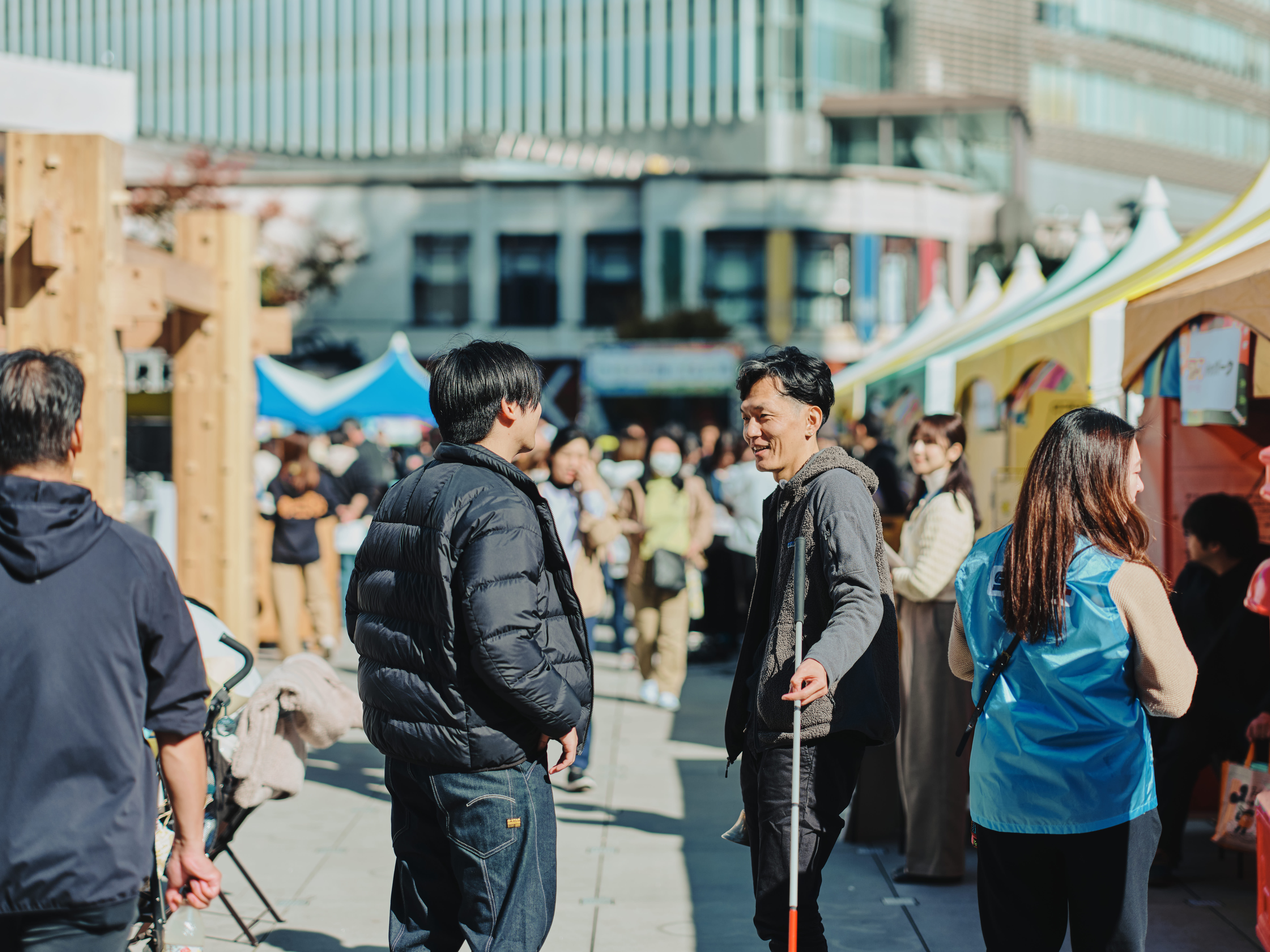 ALTはじまり:TBS主催のイベント「地球を笑顔にする広場2025秋」の屋外会場で、多くの来場者が行き交う中、黒いダウンジャケットを着た高橋さんと、グレーのフリースを着た石井さんが会話しながら立っている。テントが並ぶ明るい雰囲気の通り。ALTおわり
