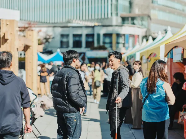 ALTはじまり:TBS主催のイベント「地球を笑顔にする広場2025秋」の屋外会場で、多くの来場者が行き交う中、黒いダウンジャケットを着た高橋さんと、グレーのフリースを着た石井さんが会話しながら立っている。テントが並ぶ明るい雰囲気の通り。ALTおわり