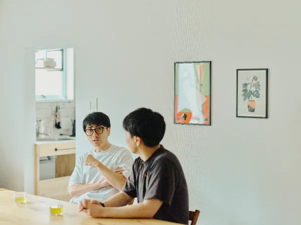 ALTはじまり: 白いTシャツを着た犬飼さんが身振り手振りを交えながら話し、大塚さんがそれを聞いている様子。テーブルにはお茶が置かれ、リラックスした雰囲気で話しています。ALTおわり