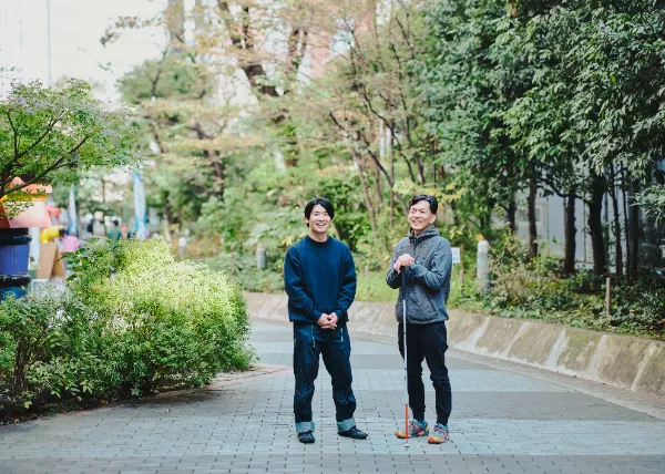 ALTはじまり:遊歩道の中央で石井さんと高橋さんが立ち、笑顔でカメラの方向を向いている。明るい日差しと緑に囲まれた写真。ALTおわり