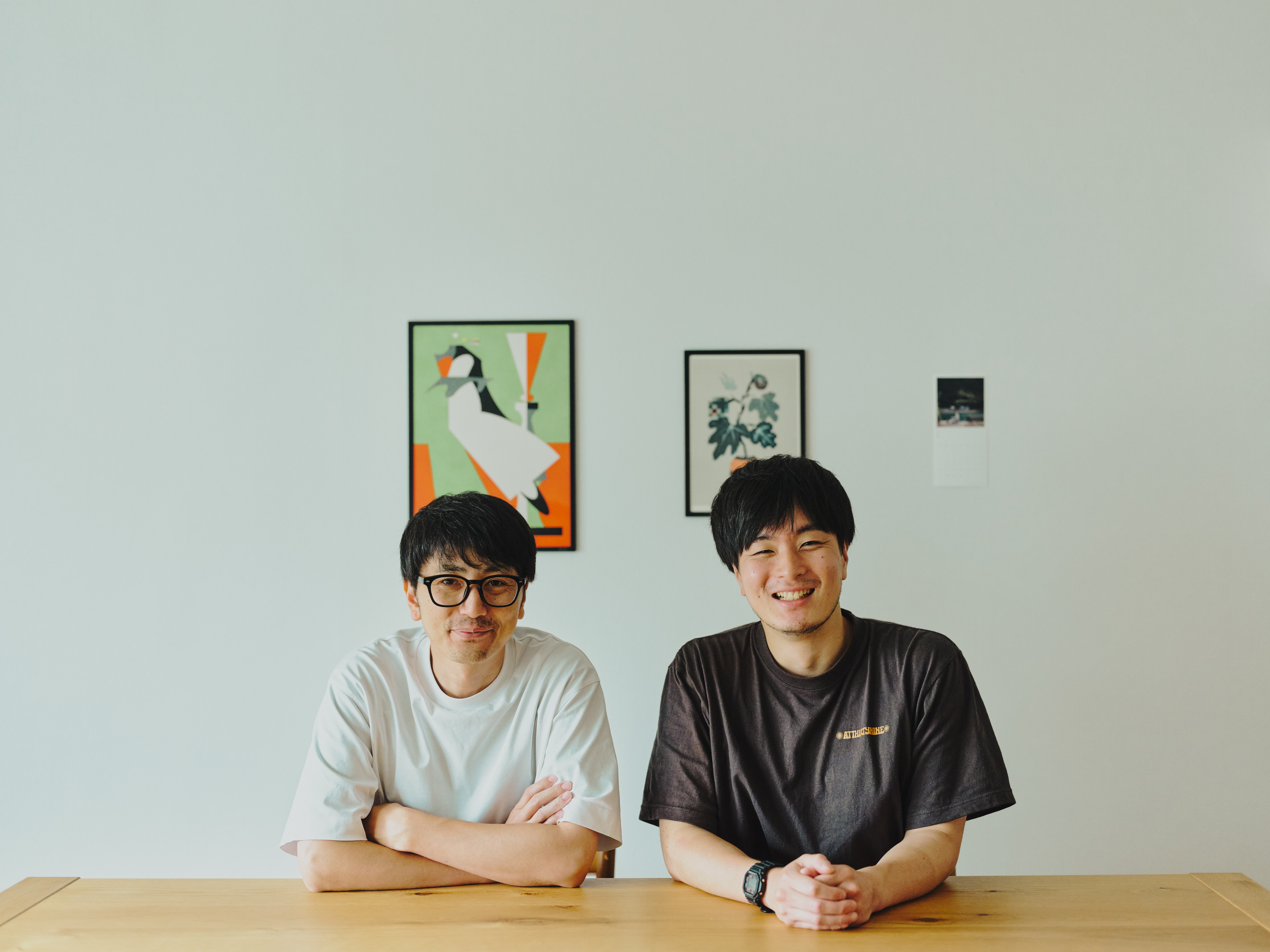 ALTはじまり:白いTシャツと眼鏡をかけた犬飼さんと、茶色いTシャツを着た大塚さんが、木製のテーブルの前に並んで座り、正面を向いてにこやかに微笑んでいます。犬飼さんは腕を組み、大塚さんは両手を組んでいます。背景の白い壁には、複数のアートが飾られています。ALTおわり