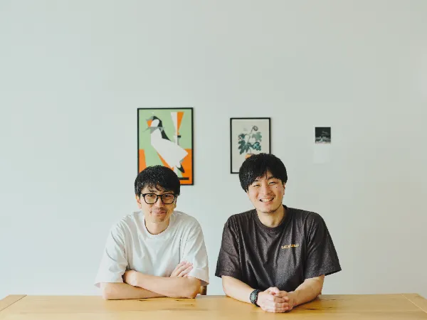 ALTはじまり:白いTシャツと眼鏡をかけた犬飼さんと、茶色いTシャツを着た大塚さんが、木製のテーブルの前に並んで座り、正面を向いてにこやかに微笑んでいます。犬飼さんは腕を組み、大塚さんは両手を組んでいます。背景の白い壁には、複数のアートが飾られています。ALTおわり