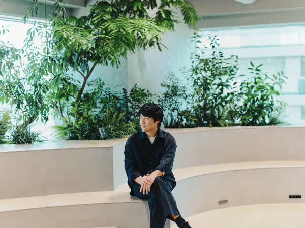 ALTはじまり:植栽に囲まれたスペースで、リラックスした様子で座り、遠くを見つめる兼原さん。場所は、25年9月にオープンしたばかりの丸井グループ本社内にある「共創LOUNGE」。世界的なデザインオフィスnendoが監修した。ALTおわり