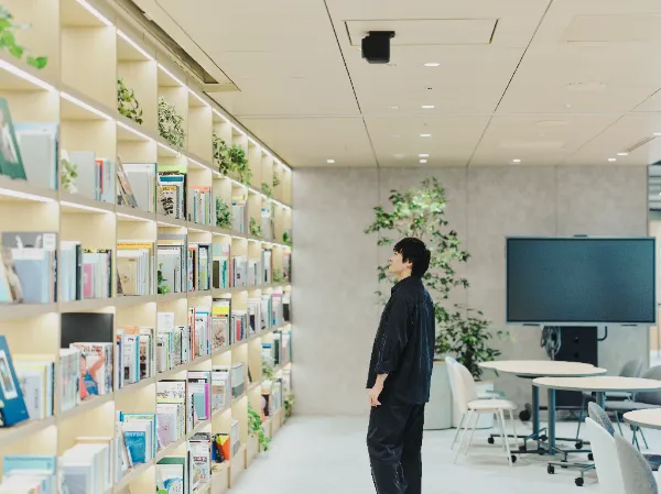 ALTはじまり: ラウンジに置かれた壁一面の本棚の前で、書籍を見上げる兼原さん。対話の一場面。ALTおわり