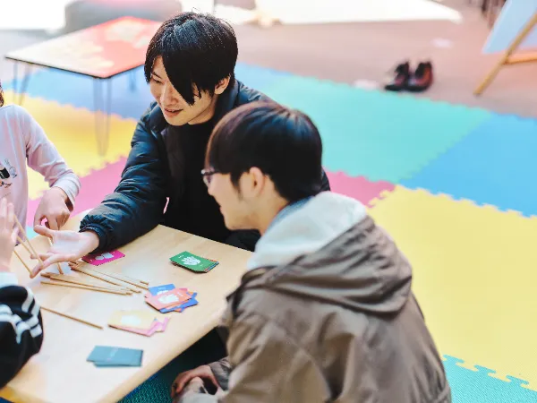ALTはじまり:カラフルなマットが敷かれたスペースで、テーブルを囲んで遊ぶ高橋さんと参加者の様子。高橋さんが発明した「YUBIBO」を使って、大人も子供も楽しそうに交流している。 ALTおわり