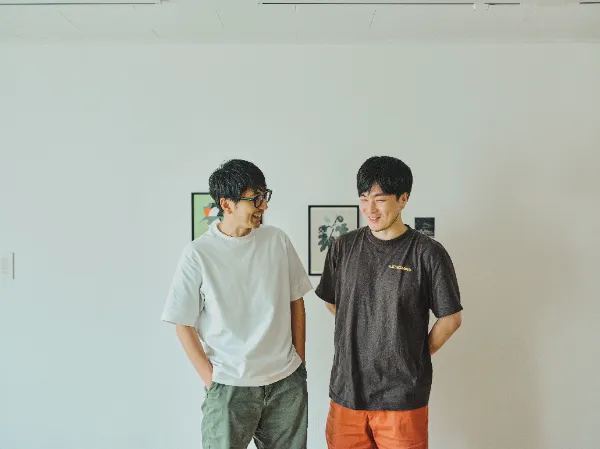 ALTはじまり: 白いTシャツを着てカーキ色のパンツを履いた犬飼さんと、茶色いTシャツを着てオレンジ色のパンツを履いた大塚さんが、横に並んで互いに顔を見合わせ、笑いながら話しています。二人ともリラックスした立ち姿で、大塚さんは両手を後ろに組んでいます。背景の白い壁にはアートが飾られています。ALTおわり