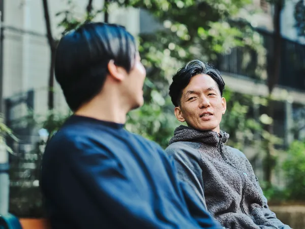 ALTはじまり:ベンチに座って向かい合い、会話をしている2人。手前に座る高橋さんはぼんやりと映り、奥に座る石井さんが話している様子がはっきり写っている。ALTおわり