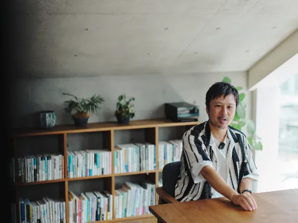 ALTはじまり: ベイジのオフィスで、木製の棚と本を背景に座り、穏やかに話すベイジ代表の枌谷力さん。ALTおわり