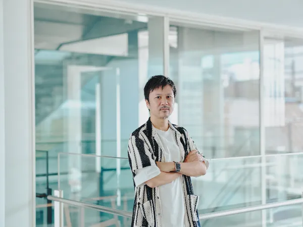 ALTはじまり:白いTシャツの上に黒と白のストライプ柄の半袖シャツを着たベイジ代表の枌谷力さんが、オフィスビル内のガラス張りの空間で、腕を組んでカメラをまっすぐ見つめている。ALTおわり