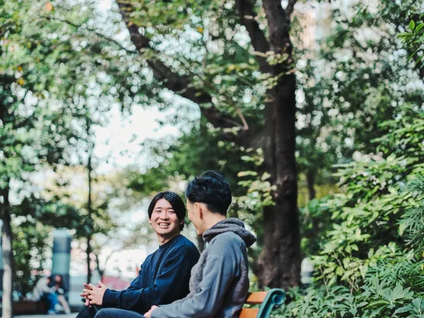 ALTはじまり:木々に囲まれた屋外で、ベンチに座った石井さんと高橋さんが向かい合って微笑んでいる。背景には公園を歩く人たちが小さく映っている。ALTおわり
