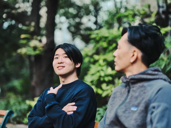 ALTはじまり:木陰のベンチで、奥に座る高橋さんが腕を組みながら少し上を見上げている。手前に座る石井さんは横向きに写っており、穏やかな時間の流れを感じる。ALTおわり