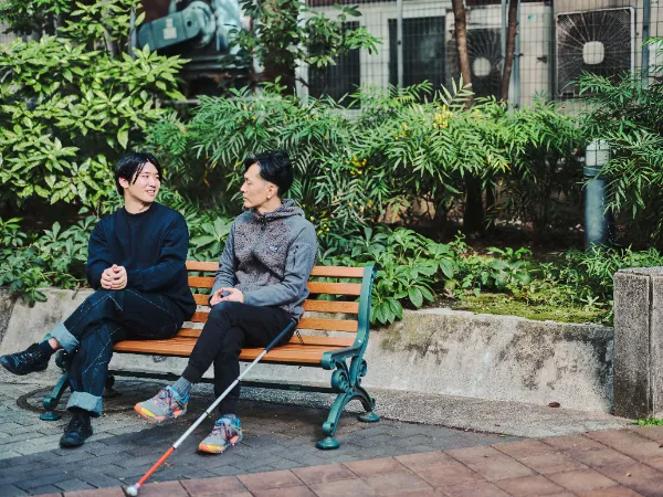 ALTはじまり:公園のベンチで向かい合って話す石井さんと高橋さん。自然の緑に囲まれた落ち着いた雰囲気の一枚。ALTおわり