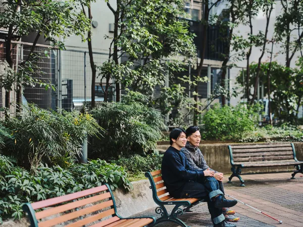 ALTはじまり:緑に囲まれたベンチで、石井さんと高橋さんが並んで座り会話している。柔らかな日差しの中、落ち着いた雰囲気の写真。ALTおわり