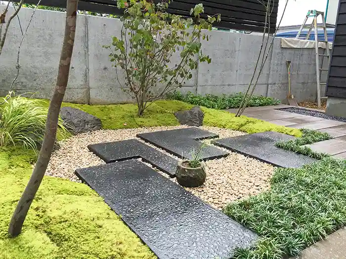 苔庭のアップ。砂利の中に配置された黒い平板のステップと、石の置物、周囲を彩るタマリュウや植栽。