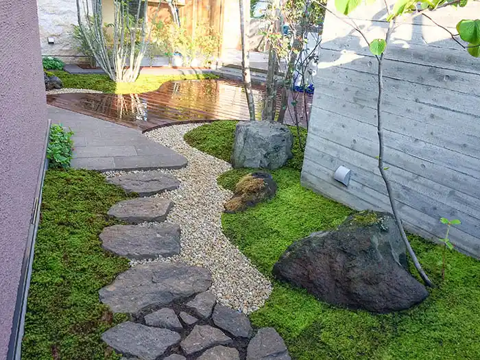 コンクリート打ち放しの壁と苔庭のコントラスト。砂利の小道が曲線を描き、大きな景石が配置された、静寂を感じさせる和風モダンな庭園風景。