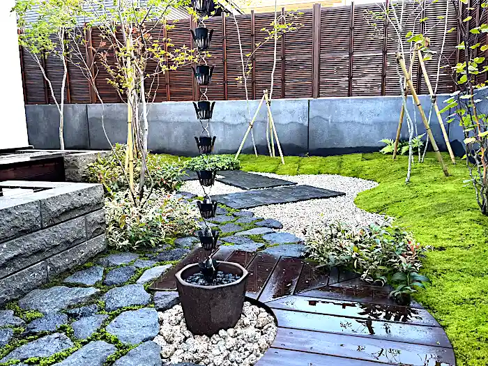 庭の中心に配置された鎖樋と水受け。背景には苔庭と白砂利、モダンなフェンスが広がる静寂な和の空間。