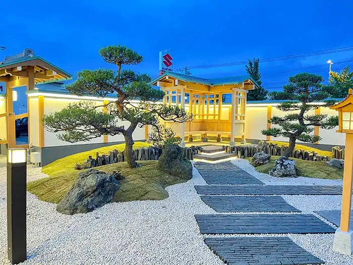 ライトアップされた夜の日本庭園。手入れされた松の木、白い砂利、モダンな長方形の石畳の小道、奥には柔らかな光を放つ木製の東屋と外灯が配置されている。