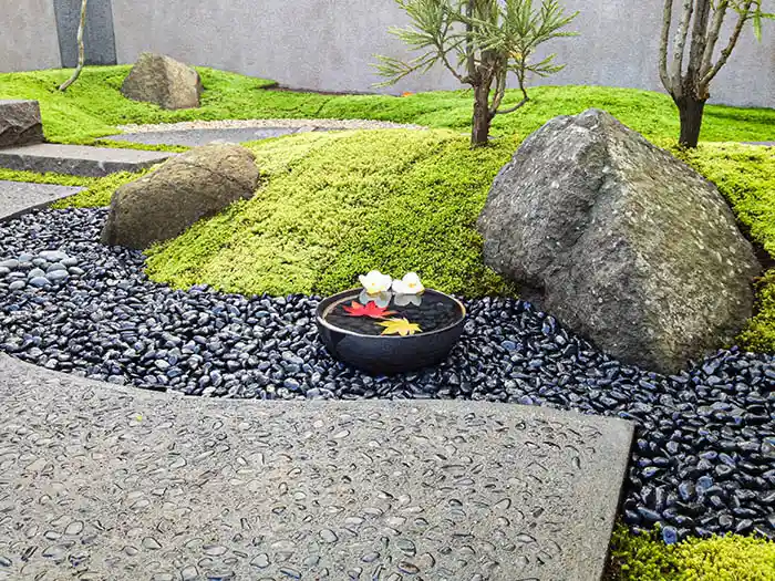 黒い玉石の流れに置かれた水鉢のアップ。水面には赤いもみじと白い花が浮かび、静寂な和の雰囲気を感じさせるディテール。