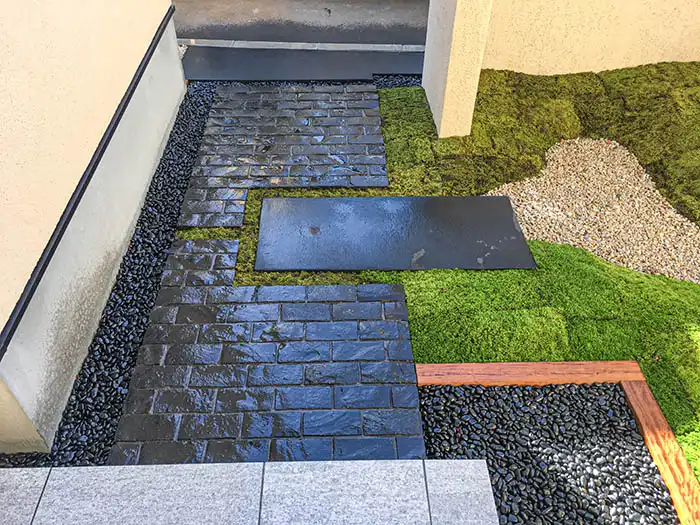 坪庭の俯瞰ショット。規則正しく並んだ黒い長方形の石畳と、柔らかな曲線を描く苔の築山、その境界を彩る白い砂利と黒い那智石の対比が美しい和モダンなアプローチ。