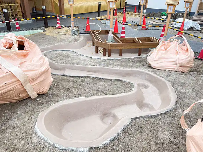 池の内側がベージュ色の防水仕上げ材でコーティングされた状態。周囲の土と馴染む色合いになり、奥には木製デッキの土台が見える。