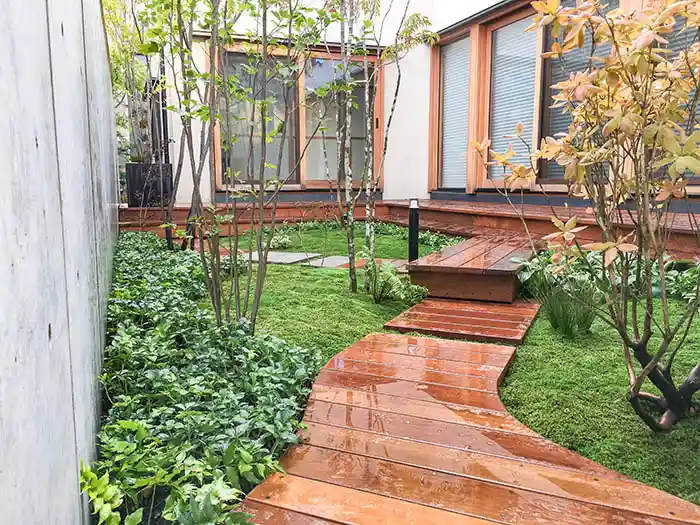 濡れたウッドパスが光を反射する、雨の日も美しい苔庭。高低差をつけたデッキと植栽が、プライバシーを守りつつ開放感を演出。
