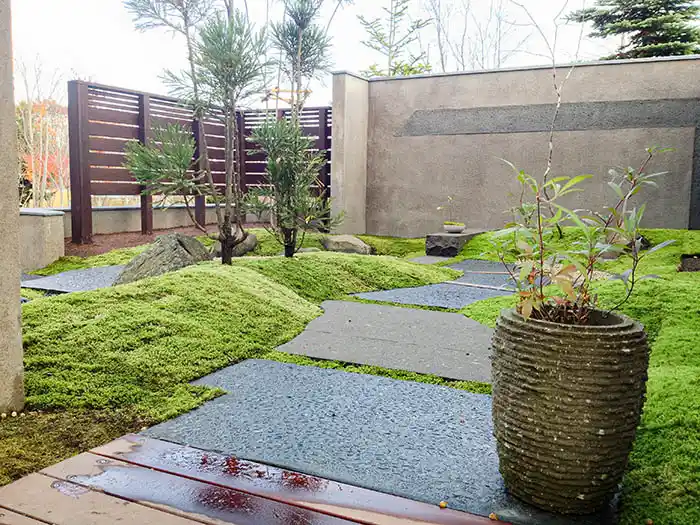 ウッドデッキから続く石畳の小道。手前には年季の入った石造りの鉢が置かれ、背景にはモダンな佐官壁と木製のフェンスがある。