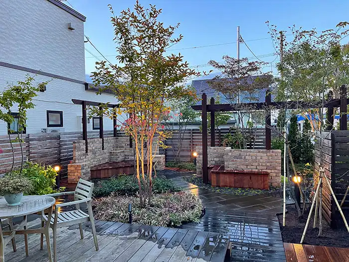 掃き出し窓前のウッドデッキテラスから見た庭の全景。テーブルセットが置かれ、シンボルツリーが夜のライトに照らされている様子。