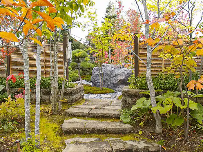 低い石垣の門を通り、庭園内部へと続く平らな石畳と、黄色や赤に色づいたモミジの風景。