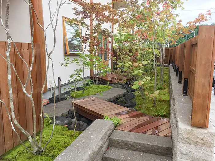 完成した庭。苔が生い茂り、木々の間を抜けるように石組みの流れがある。手前には木製のデッキと階段、奥には建物へ続く小道が見える。